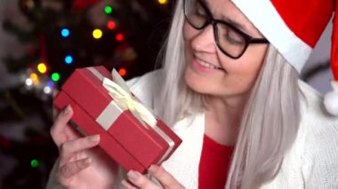 Mutlu kadın Noel ağacı için hediye kutusunu açacağı için heyecanlı. Noel Baba şapkalı kız kameraya bakıyor ve el sallıyor. Noel hediyesi. Aile geleneği. 4k video