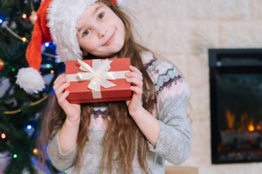 Noel Baba şapkalı mutlu kız içinde ne olduğunu öğrenmek için hediye kutusu tutuyor. Çocuk Noel ışıklarıyla süslenmiş Noel ağacından önce evde yatıyor. Tatil ve kutlama konsepti