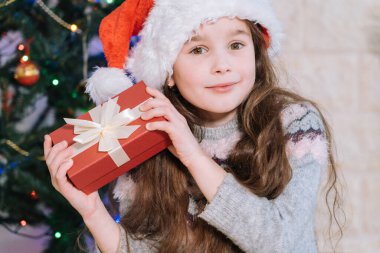 Noel Baba şapkalı mutlu kız içinde ne olduğunu öğrenmek için hediye kutusu tutuyor. Çocuk Noel ışıklarıyla süslenmiş Noel ağacından önce evde yatıyor. Tatil ve kutlama konsepti