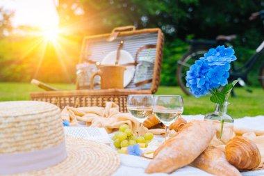 Güneşli bir günde yaz pikniği. Ekmek, meyve, buket ortanca çiçeği, bardak şarabı, hasır şapka, kitap ve ukulele. Çimlerin üzerinde piknik sepeti, yiyecek ve beyaz örgü battaniyenin üzerinde içki. Seçici odak.