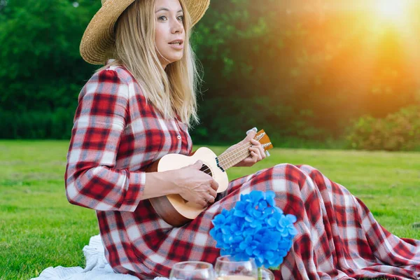 Girl in red checkered dress and hat sitting on white knit picnic blanket plays ukulele and drinking wine. Summer picnic on sunny day with bread, fruit, bouquet hydrangea flowers. — Stock Photo, Image