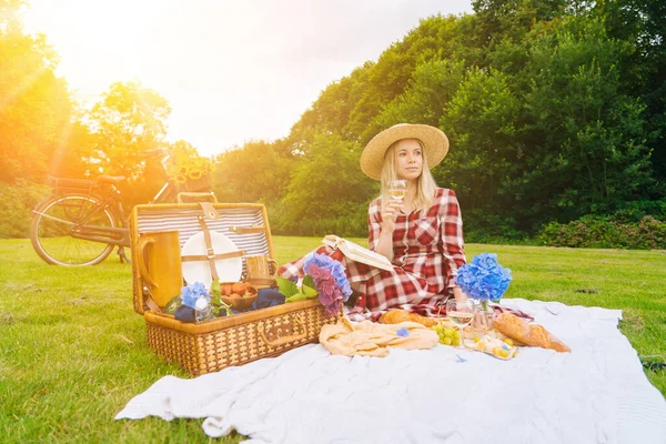 Girl in red checkered dress and hat sitting on white knit picnic blanket reading book and drinking wine. Summer picnic on sunny day with bread, fruit, bouquet hydrangea flowers. Selective focus. — Stock Photo, Image