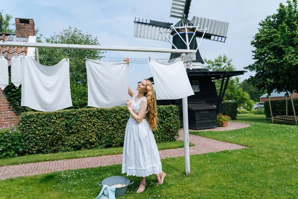 Mujer vestida con el tradicional vestido holandés zapatos de madera zuecos amarillos colgando Ropa en el tendedero exterior. lavandería secado al aire libre Países Bajos molino de viento de fondo en el día ventoso sol. estilo vintage retro Fotos De Stock