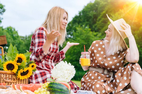 Friends is making picnic outdoor. Laughing girls sitting on white knit picnic blanket drinking wine. Happy young women talking, smiling outdoors on Summer picnic on sunny day at park.