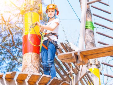 Turuncu kasklı çocuk orman macerası parkında ağaçlara tırmanıyor. Kız, yaz parkında halat kablolarında ve yüksek asma köprüde yürüyor. Ekstrem spor malzemesi miğferi ve karabina.
