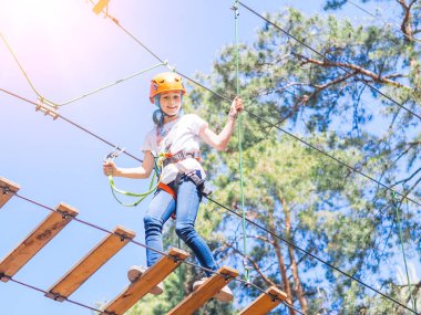 Turuncu kasklı çocuk orman macerası parkında ağaçlara tırmanıyor. Kız, yaz parkında halat kablolarında ve yüksek asma köprüde yürüyor. Ekstrem spor malzemesi miğferi ve karabina.
