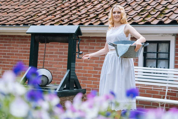 Woman in white dress holding basin near ancient artesian draw-well in village. Colorful violet flowers on foreground. Girl gets water from rural well in summer sunny day for washing. — Stock Photo, Image