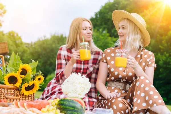 Friends is making picnic outdoor. Laughing girls sitting on white knit picnic blanket drinking wine. Happy young women talking, smiling outdoors on Summer picnic on sunny day at park.