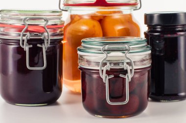 Beyaz bir zemin üzerinde düzenlenmiş çilek reçeli, şeftali kompostosu, erik yağı ve böğürtlen reçeli gibi ev yapımı meyve reçelleri, yemek, tarif veya mutfak temalı tasarımlar için idealdir.