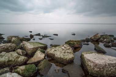 Yeşil alglerle kaplı taşlar ve kayalar. Pürüzsüz temiz su. Arkaplanda bulutlu bir gökyüzü ve hafif bir sis.