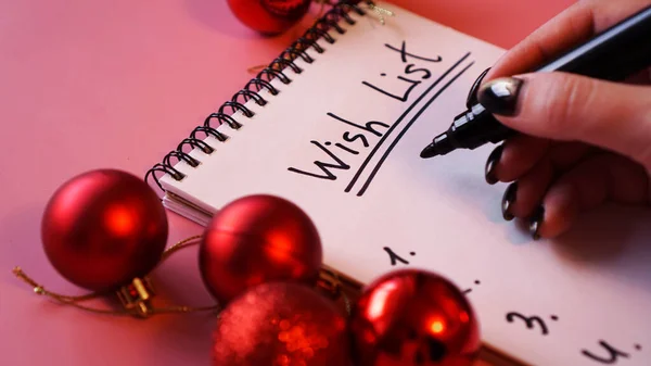 Frau schreibt ihre Wunschliste. Festliches Design auf rosa Hintergrund mit roten Kugeln — Stockfoto