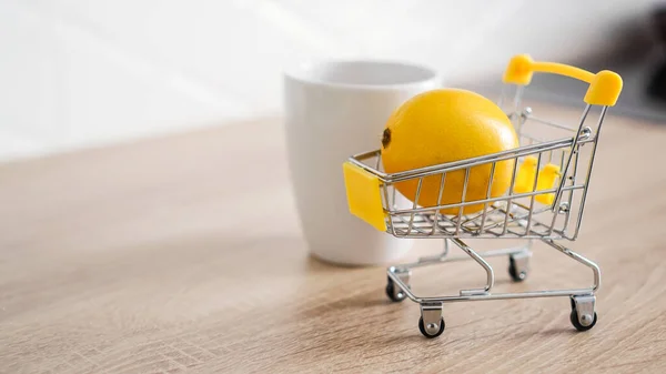 Citron v malém nákupním vozíku na kuchyňském stole. Nedaleko je bílý hrnek čaje — Stock fotografie
