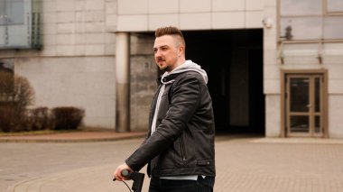 Gri şehir binalarının arka planında elektrikli scooter kullanan uzun boylu bir adam.