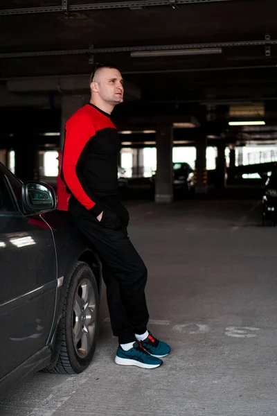 Um jovem de fato de treino está ao lado de um carro num estacionamento subterrâneo. — Fotografia de Stock