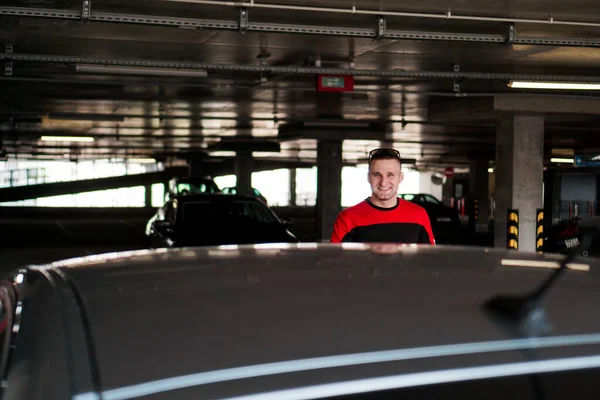 A young man in a tracksuit stands next to a car in an underground parking lot — Stock Photo, Image