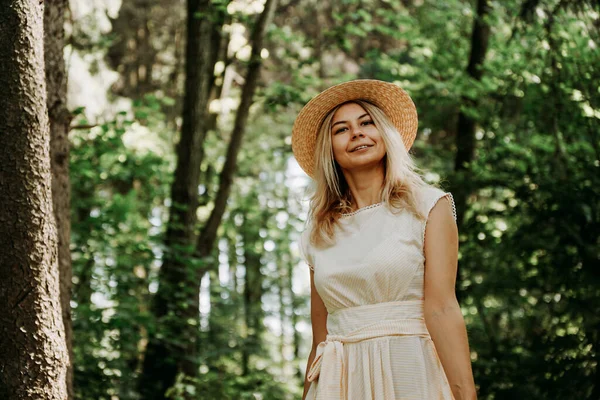 Bella giovane donna con cappello di paglia e vestito bianco in un parco verde — Foto Stock