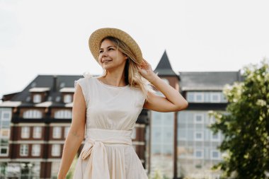 Güzel bir sarışın şehirde yürüyor. Beyaz elbiseli ve hasır şapkalı kadın.