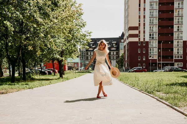 Piękna blondynka spaceruje po mieście. Kobieta w białej sukience i słomkowym kapeluszu — Zdjęcie stockowe