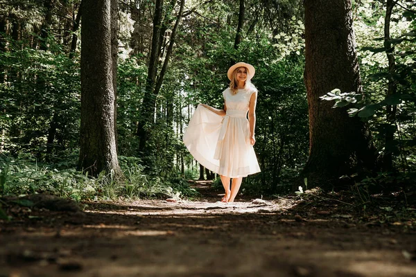 Una niña con un vestido blanco y sombrero camina a través de un parque de verano o bosque — Foto de Stock