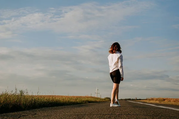 Una mujer pelirroja con una camisa blanca camina por el camino entre los campos —  Fotos de Stock