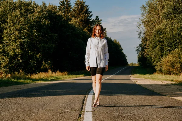 Une femme aux cheveux roux en chemise blanche marche le long de la route au milieu de la forêt — Photo