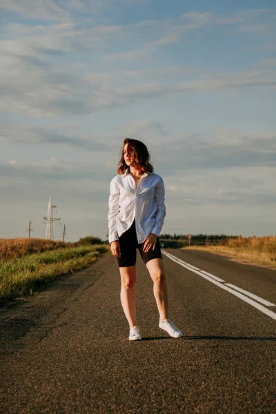 Een roodharige vrouw in een wit shirt loopt langs de weg tussen de velden — Stockfoto