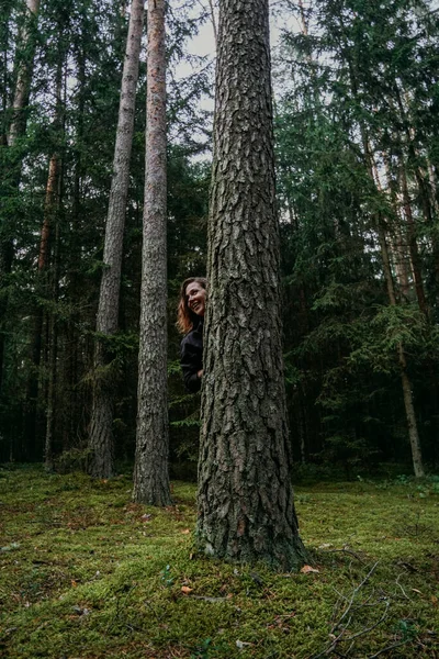 Una joven en un bosque de coníferas mira desde detrás de un árbol —  Fotos de Stock
