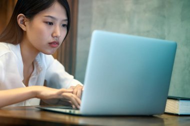 Zeki, aktif, stresli Asyalı kadın geç saatlere kadar çalışır. İşyerindeki ahşap masada dizüstü bilgisayarla çalışmaya konsantre olur.