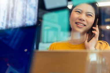 Yaratıcı ajans Asyalı kadın günlük kıyafet sohbeti. Akıllı telefon serbest bilgisayar ile birlikte çalışma alanındaki ofis alanında çalışıyor. Ofis geçmişi bulanık.