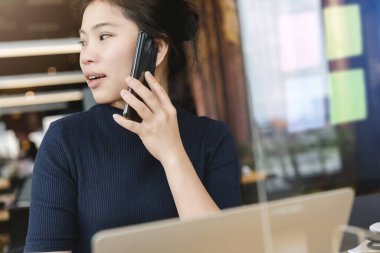 Çekici Asyalı kadın lacivert süveter koruyucu virüs maskesi takarken çalışıyor. Yeni normal yaşam tarzı için plastik bölme sosyal mesafesi olan birlikte çalışma alanında çalışıyor.