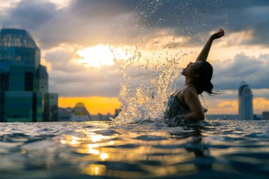 Asyalı bir kadının siyah silueti yaz tatilinde su sıçratıyor. Sonsuz yüzme havuzunda dinleniyor. Mavi deniz batımı manzaralı, gökyüzü manzaralı şehir merkezinde sağlıklı bir yaşam tarzı.
