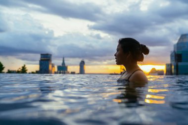 Asyalı bir kadının siyah silueti yaz tatilinde su sıçratıyor. Sonsuz yüzme havuzunda dinleniyor. Mavi deniz batımı manzaralı, gökyüzü manzaralı şehir merkezinde sağlıklı bir yaşam tarzı.