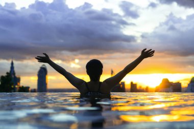 Asyalı bir kadının siyah silueti yaz tatilinde su sıçratıyor. Sonsuz yüzme havuzunda dinleniyor. Mavi deniz batımı manzaralı, gökyüzü manzaralı şehir merkezinde sağlıklı bir yaşam tarzı.