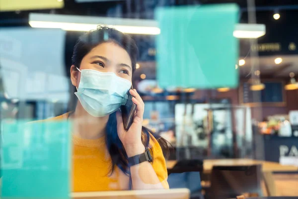 creative agency asian female casual cloth and face mask protection conversation with smartphone freelance working with laptop at coworking area office space with freshness with blur office background