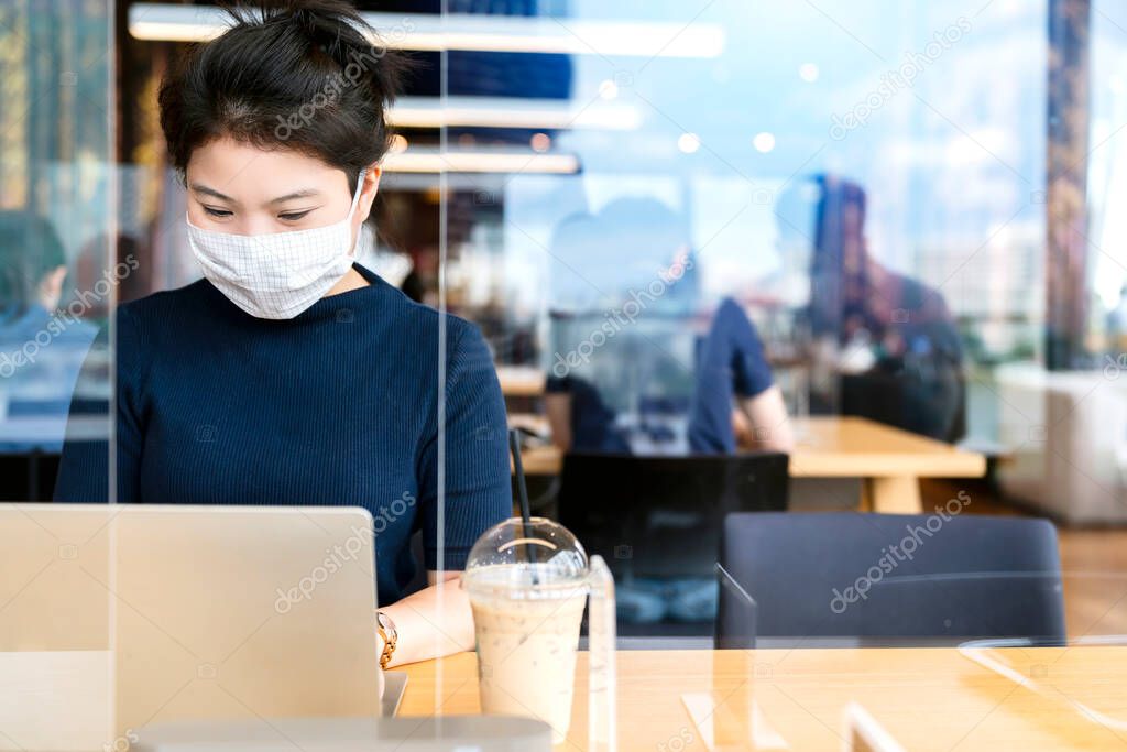 attractive asian female navy blue sweater working while wearing protective virus mask working in co woking space that have plastic partition social distancing for seat new normal lifestyle