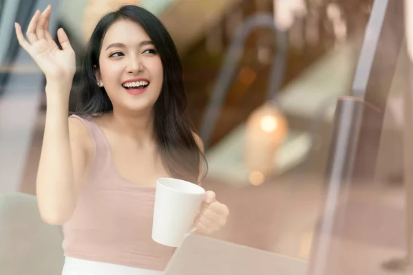 attractive cheerful asian female adult gesture hand wave to say hi to her friend walking in cafe restaurant,asian woman hand hold coffee cup relax casual in cafe shot through window glass