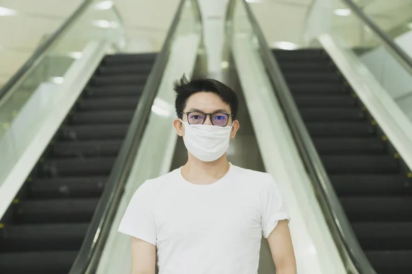 young asiang glasses male wear protecting white mask to avoid corona virus or covid 19 spread ,asian man stand next to escalator urban lifestyle