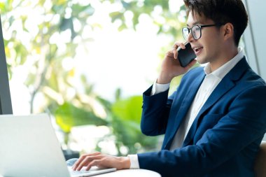 yaratıcı ajans akıllı gözlükler Asyalı erkek resmi kumaş konuşma akıllı telefon serbest çalışma alanı ofis alanında laptop ile çalışıyor bulanık bahçe ağacı arka planı ile tazelik