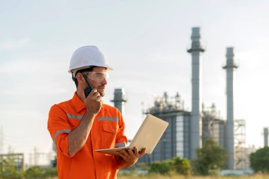 Beyaz erkek, mühendis, endüstriyel işçiler güvenli üniforma giyiyorlar. Telsiz ve dizüstü bilgisayarla elektrik santralinin arka planında araştırma yapıyorlar.