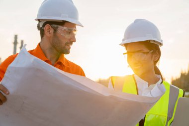 Asyalı kadın mühendis, beyaz erkek, endüstriyel işçiler güvenlik üniforması giyip telsiz ve taslak üzerinde çalışıyorlar.