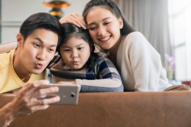 Genç mutlu Asyalı aile el resmi neşeli ve eğlenceli karantina dönemi yeni normal yaşam tarzı, anne ve baba oturma odasındaki kanepede birlikte fotoğraf çekiyorlar.