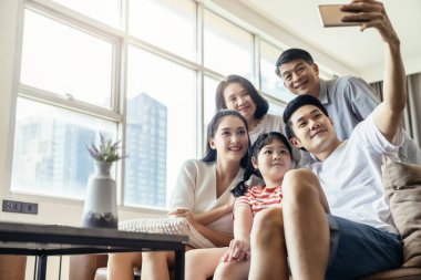 Asyalı çoklu nesil aile babası ve küçük kızı olan büyük ebeveyn oturma odasındaki koltukta oturup akıllı telefonlu selfie çekiyorlar. Evde gülümseyen ve gülen bir gülümsemeyle.