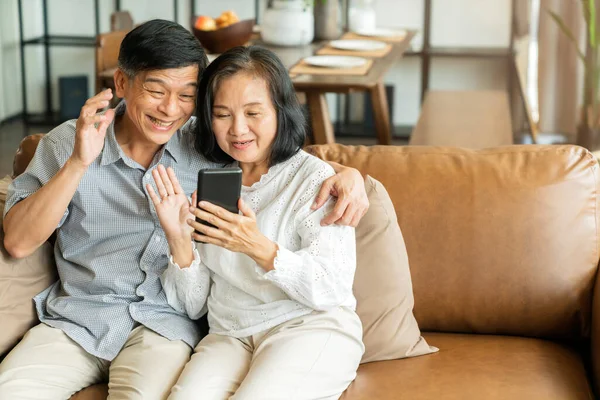 快乐的老亚洲情人夫妇手握智能手机 看着手机屏幕轻松地坐在沙发上 笑着年长的祖父母家人拥抱生活方式 — 图库照片