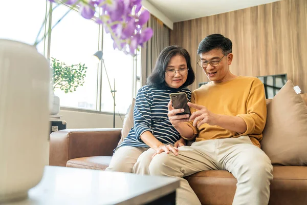 快乐的老亚洲情人夫妇手握智能手机 看着手机屏幕轻松地坐在沙发上 笑着年长的祖父母家人拥抱生活方式 — 图库照片