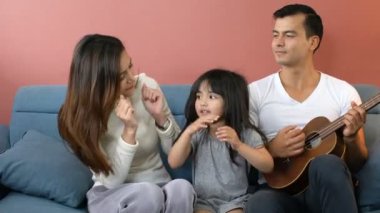 Mutlu bir aile kızı ve ebeveyni olan bir kız çocuğu oturma odası ilişkisindeki kanepede sevgi ve neşeyle oynuyorlar.