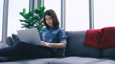 Güzel, zeki Asyalı kadın oturma odasının arka planında dizüstü bilgisayar dükkanıyla rahatla.