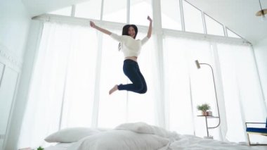 Asyalı kadının yavaş çekimde yatak odasındaki beyaz yatağın üzerinden atlaması enerji ve başarı dolu yaşam tarzı fikirleri kavramı.