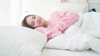 Çok güzel bir Asyalı kadın beyaz yatak odasında huzur içinde uyur. Pencereden güneş ışığı gelir.