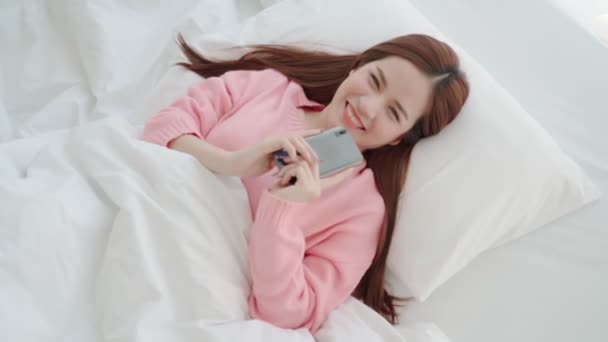 Mooie lange haar aziatische vrouw mooie glimlach genieten smartphone chat na wakker ochtend weekend op bed slaapkamer interieur achtergrond — Stockvideo