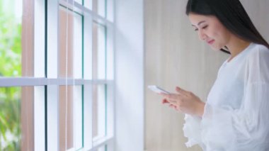 Güzel Asyalı kadın mutluluğun tadını çıkar. Pencerenin yanında akıllı telefon kullan.